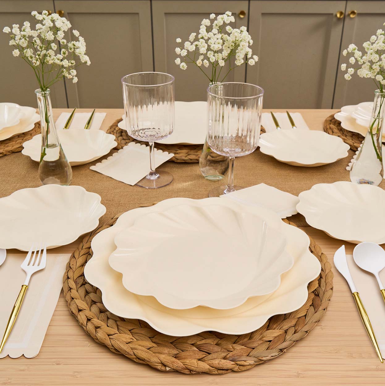 The Eco-Friendly Table Setting-Cream by Sophistiplate features cream compostable plates, gold and white cutlery, woven placemats, ribbed glasses, and white flower vases on a tan runner against a neutral backdrop.