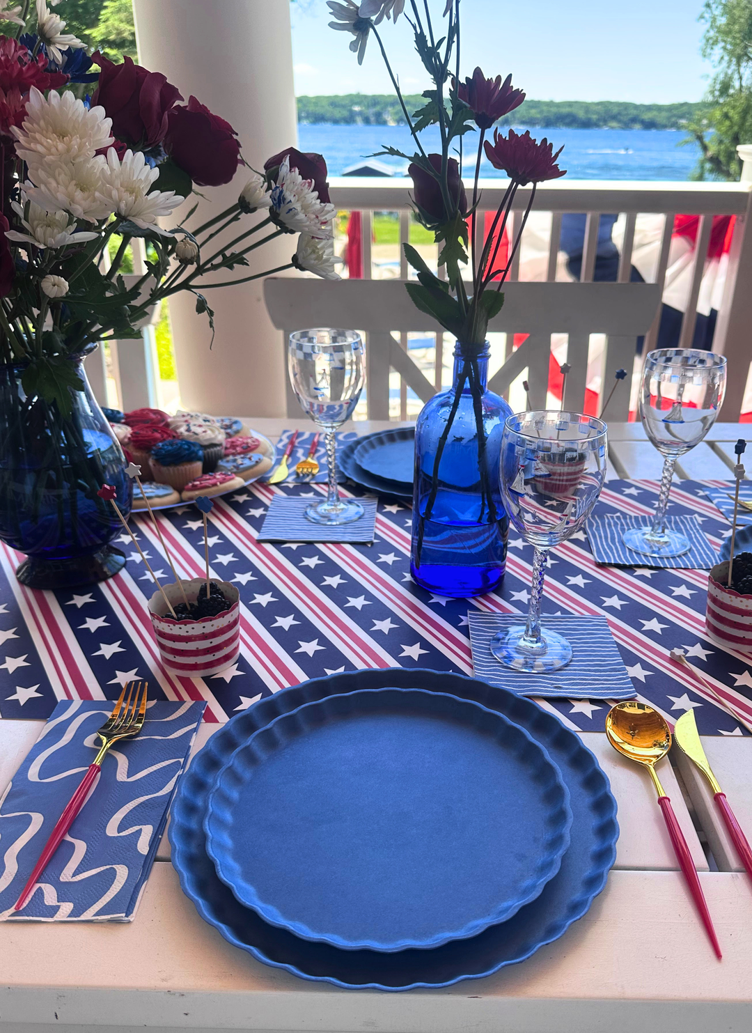 Celebrate lakeside with an eco-friendly 4th of July Table for 16+ in French Blue from Sophistiplate LLC—featuring blue plates, gold and red cutlery, flag napkins, blue vases, and a festive party pack for glasses and desserts.
