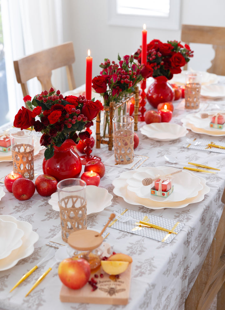 A festive table is set with the Sophistiplate LLC Rosh Tablescape for 16 Guests: Eco Cream Honeybee, featuring elegant white compostable plates, gold cutlery, red candles, vases of red flowers, apples, honey jars, and wrapped gifts on a white patterned tablecloth—creating a vibrant and inviting atmosphere.