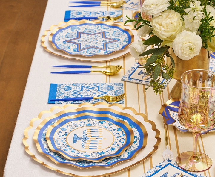 Create a festive tablescape with the Hanukkah Toile Table Setting by Sophistiplate, featuring elegant blue and white disposable plates, gold cutlery, coordinating blue napkins, a white floral centerpiece, and gold-accented glassware set atop a crisp white tablecloth.
