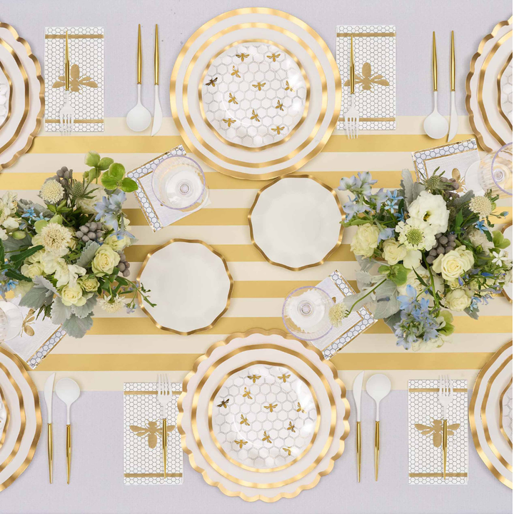 A table set for six with Sophistiplate Honeybee White Wavy Paper Dinner Plates, gold utensils, bee-themed napkins, and floral centerpieces on a gold and white striped runner.
