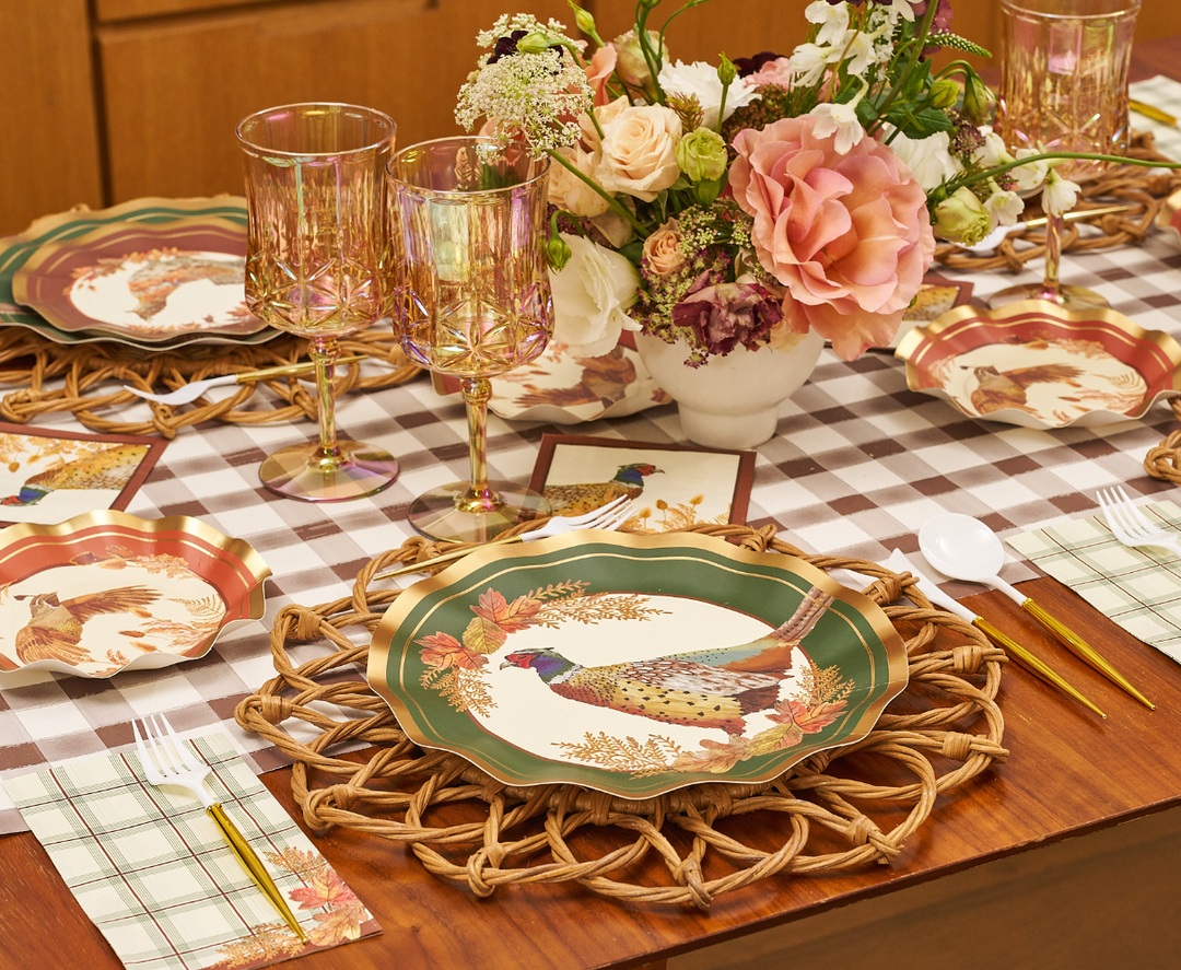 A table is set with pheasant-themed plates, gold utensils, Sophistiplate Woodland Harvest Guest Towels as napkins, pink glassware, and a floral centerpiece on a brown and white checked tablecloth.