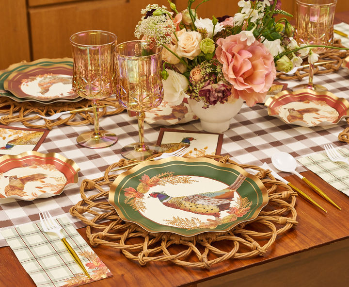 A table is set with pheasant-themed plates, gold utensils, Sophistiplate Woodland Harvest Guest Towels as napkins, pink glassware, and a floral centerpiece on a brown and white checked tablecloth.