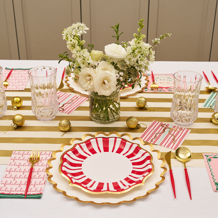 A festive table setting featuring Sophistiplate’s Tis the Season Dinner Plates (8-pack) with red and white stripes, gold cutlery, clear glasses, holiday napkins, gold ornaments, and a white floral centerpiece atop a gold-striped runner.