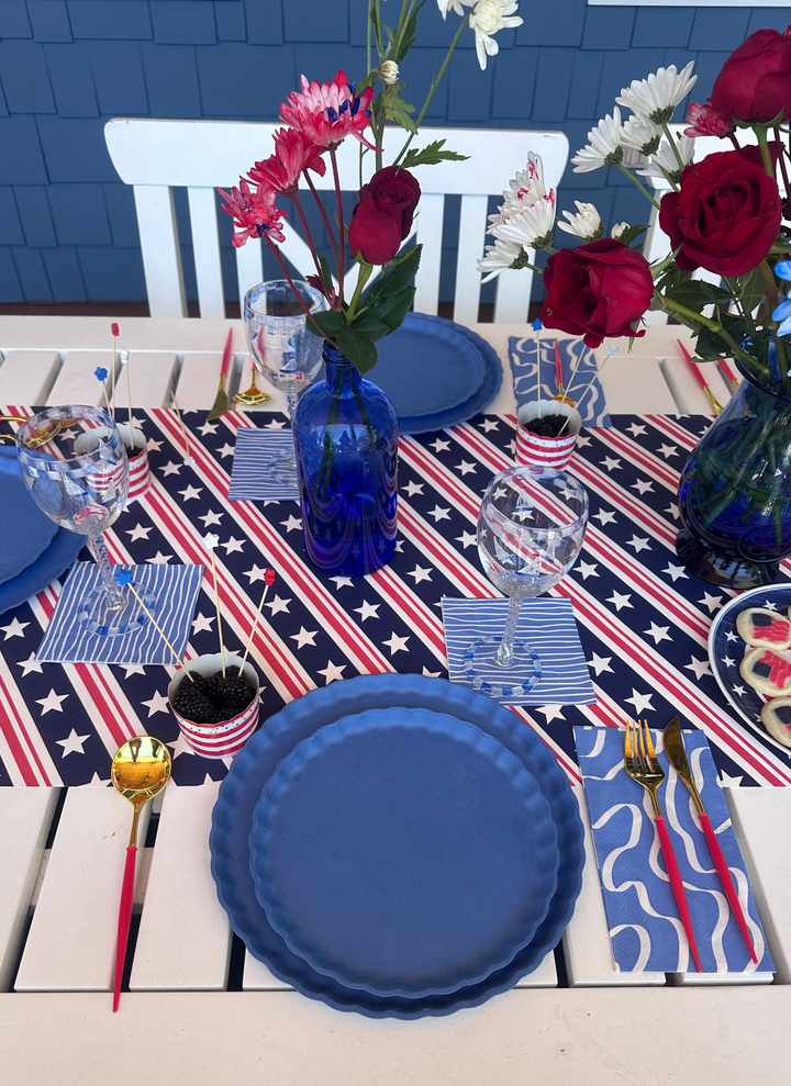 A festive 4th of July table by Sophistiplate LLC features French Blue eco-friendly plates, gold cutlery, American flag decor, red-white-blue floral vases, a patriotic runner, glasses, and sprinkle-filled bowls for 16+ guests.