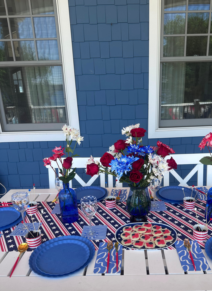 The 4th of July Table for 16+ in French Blue by Sophistiplate LLC features compostable bamboo plates, American flag napkins, a star-patterned tablecloth, and festive flowers—perfect for patriotic outdoor gatherings.