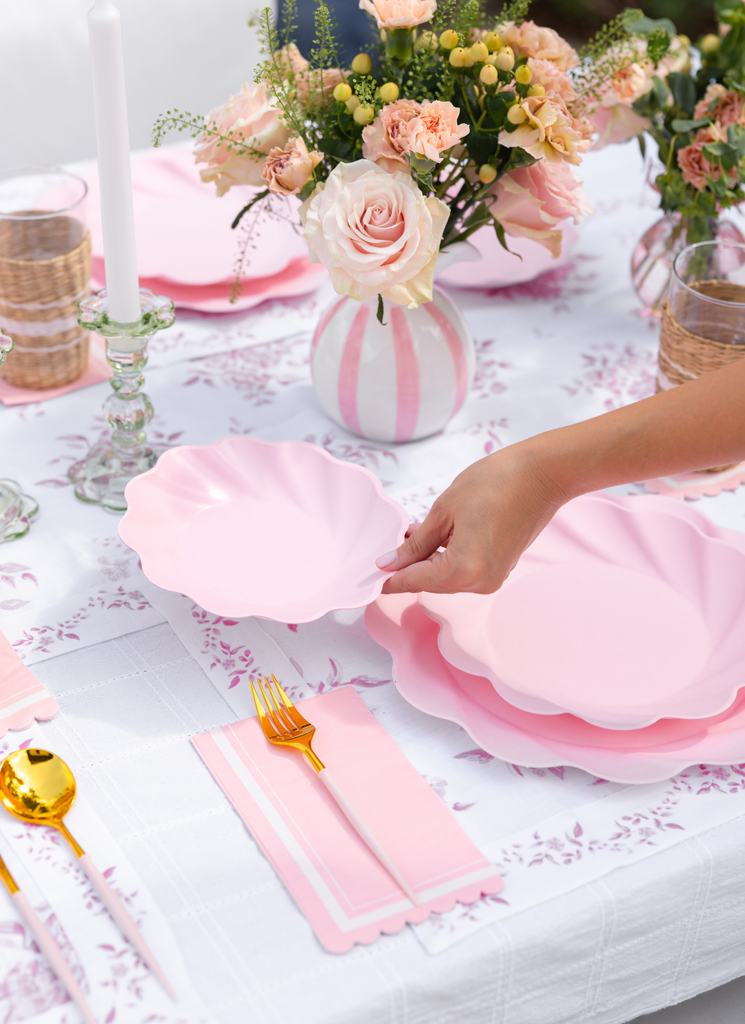 A hand sets a blush, scalloped Eco-Friendly Table Setting by Sophistiplate LLC on a floral tablecloth, surrounded by gold cutlery, pink napkins, candles, and vases of blush roses and greenery.