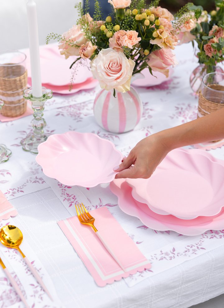A hand sets a blush, scalloped Eco-Friendly Table Setting by Sophistiplate LLC on a floral tablecloth, surrounded by gold cutlery, pink napkins, candles, and vases of blush roses and greenery.