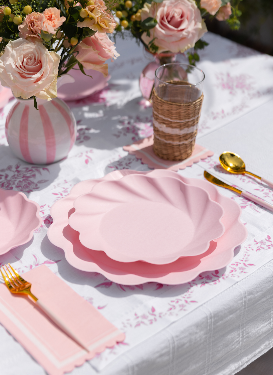 The Eco-Friendly Table Setting- Blush by Sophistiplate LLC features blush pink napkins, gold cutlery, wicker-wrapped glassware, and roses on a white floral tablecloth for a stylish, sustainable dining experience.