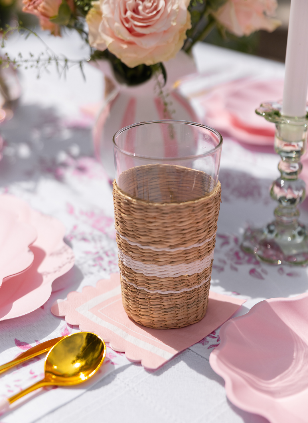 A glass in a woven holder rests on pink napkins alongside gold utensils, eco-friendly disposable plates, blush plates, and a floral centerpiece from the Eco-Friendly Table Setting- Blush by Sophistiplate LLC.