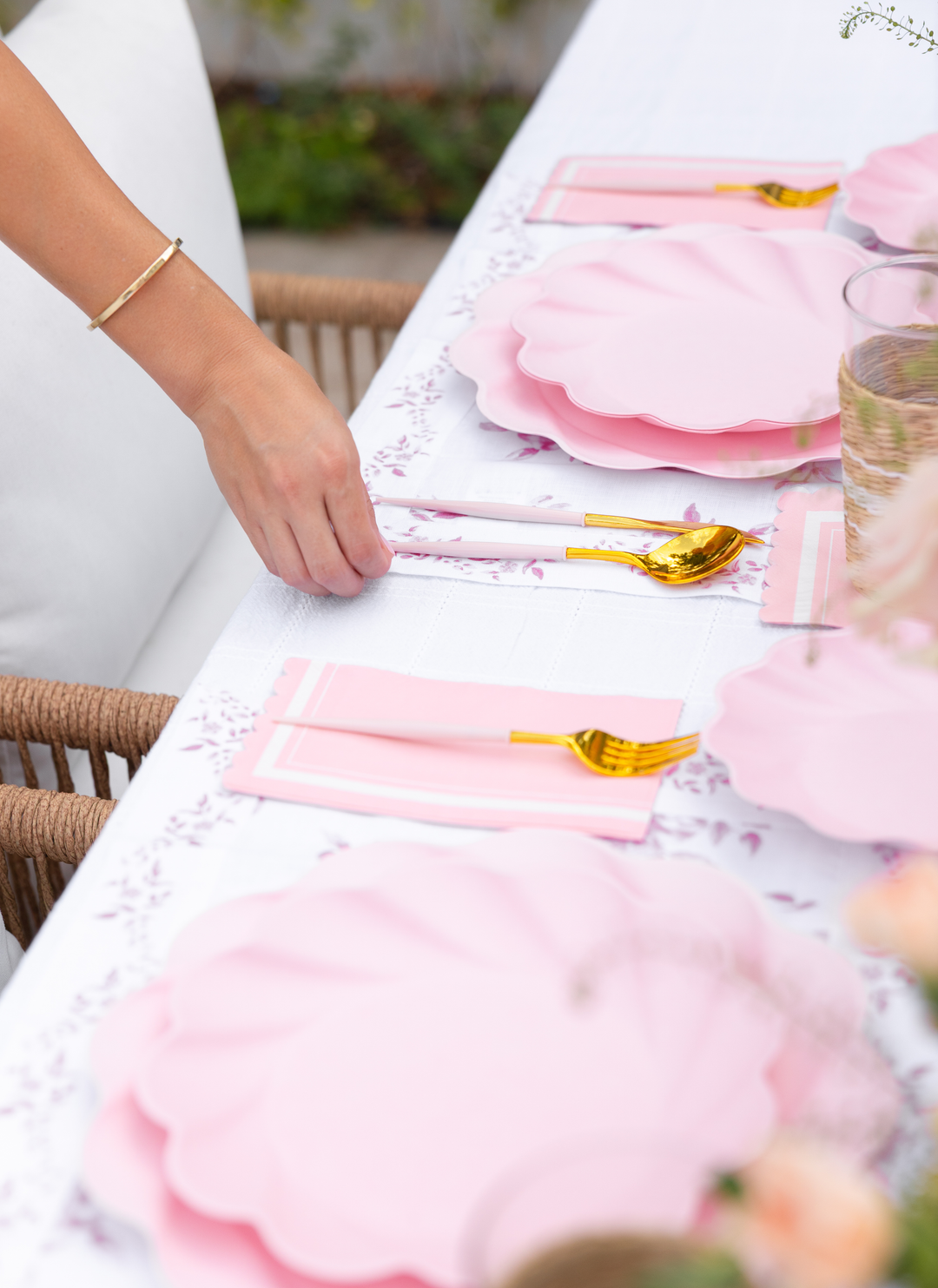 A person sets the table with Sophistiplate LLC's Eco-Friendly Table Setting- Blush, featuring pink scallop-shaped plates, gold utensils, and pink napkins on a white floral tablecloth for an elegant, festive look.