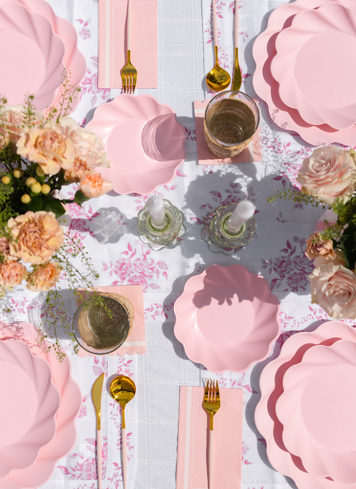 The Eco-Friendly Table Setting- Blush by Sophistiplate LLC features a table for four with pink scalloped compostable plates, gold cutlery, pink napkins, glassware, floral accents, and a white tablecloth with a pink floral design.