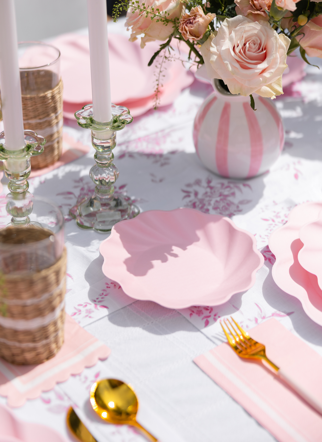 A close-up of Sophistiplate LLC's Eco-Friendly Table Setting- Blush features pink scalloped plates, gold cutlery, glassware in wicker holders, pink napkins, and a vase of pink and white flowers on a white patterned tablecloth.