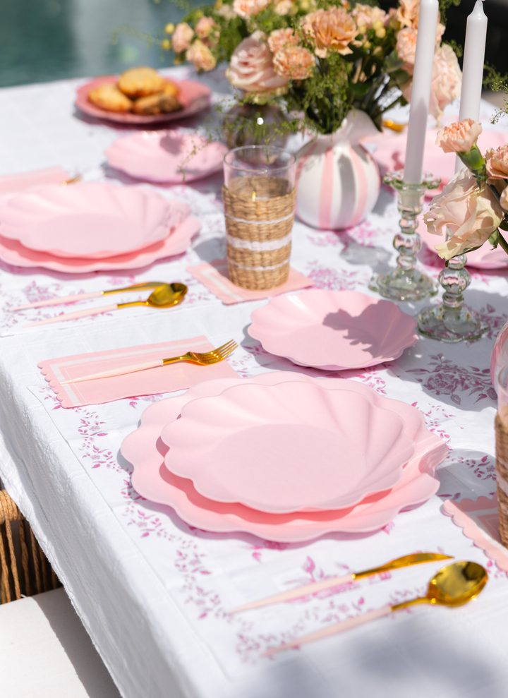 An outdoor table is set with Sophistiplate LLC’s Eco-Friendly Table Setting- Blush, featuring pink shell-shaped plates, gold utensils, floral napkins, flowers in vases, lit candles, a glass of water, and cookies on a white tablecloth.