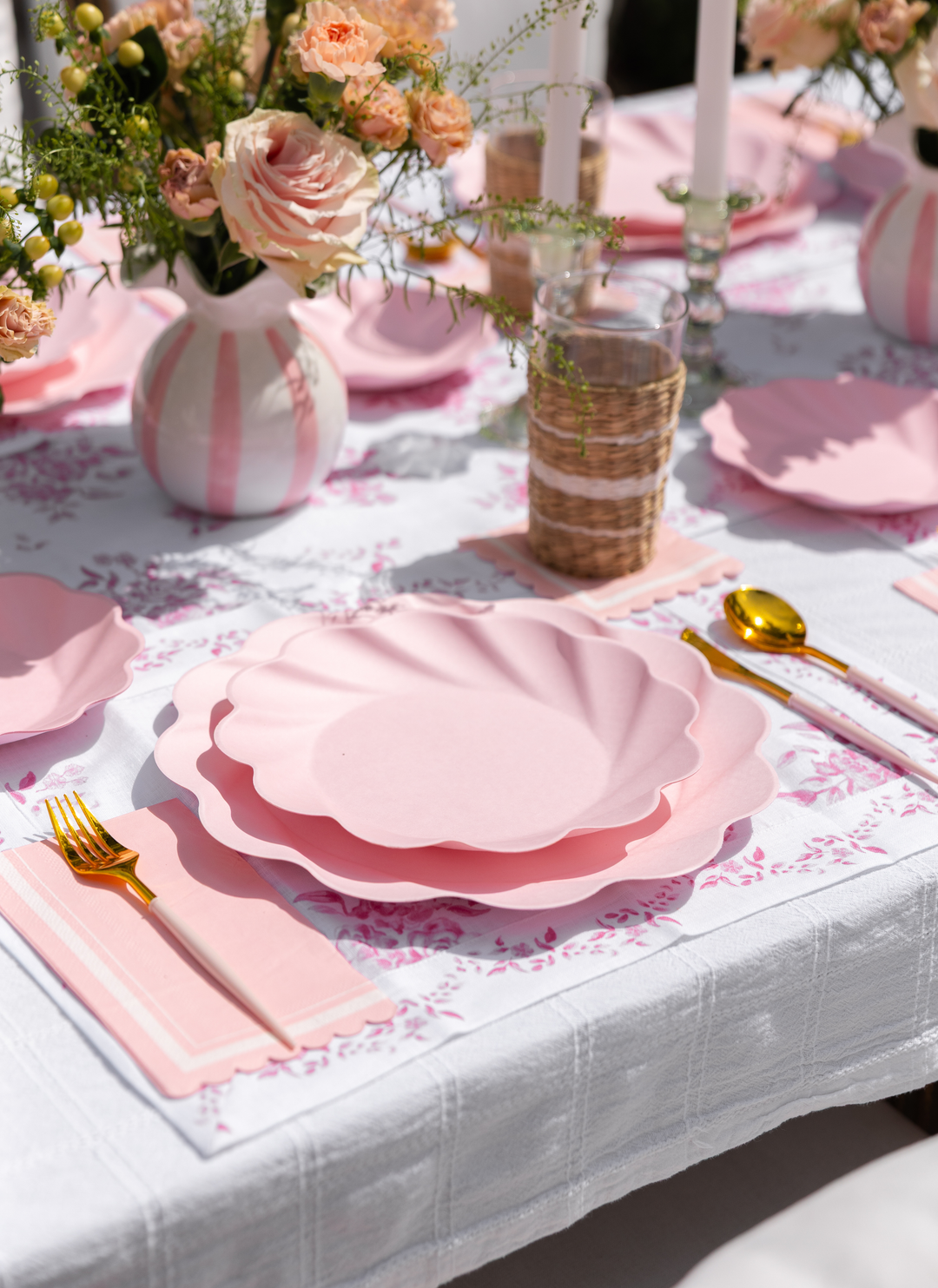 The Eco-Friendly Table Setting- Blush by Sophistiplate LLC features disposable plates, gold cutlery, woven glasses, floral napkins, and vases of pink roses on a white patterned tablecloth, with tall white candles for an elegant touch.