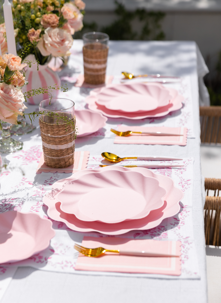 A sunlit outdoor table is set for a meal with the Eco-Friendly Table Setting- Blush by Sophistiplate LLC, featuring compostable party plates, gold utensils, pink napkins, woven glasses, and vases of blush roses on a white floral tablecloth.