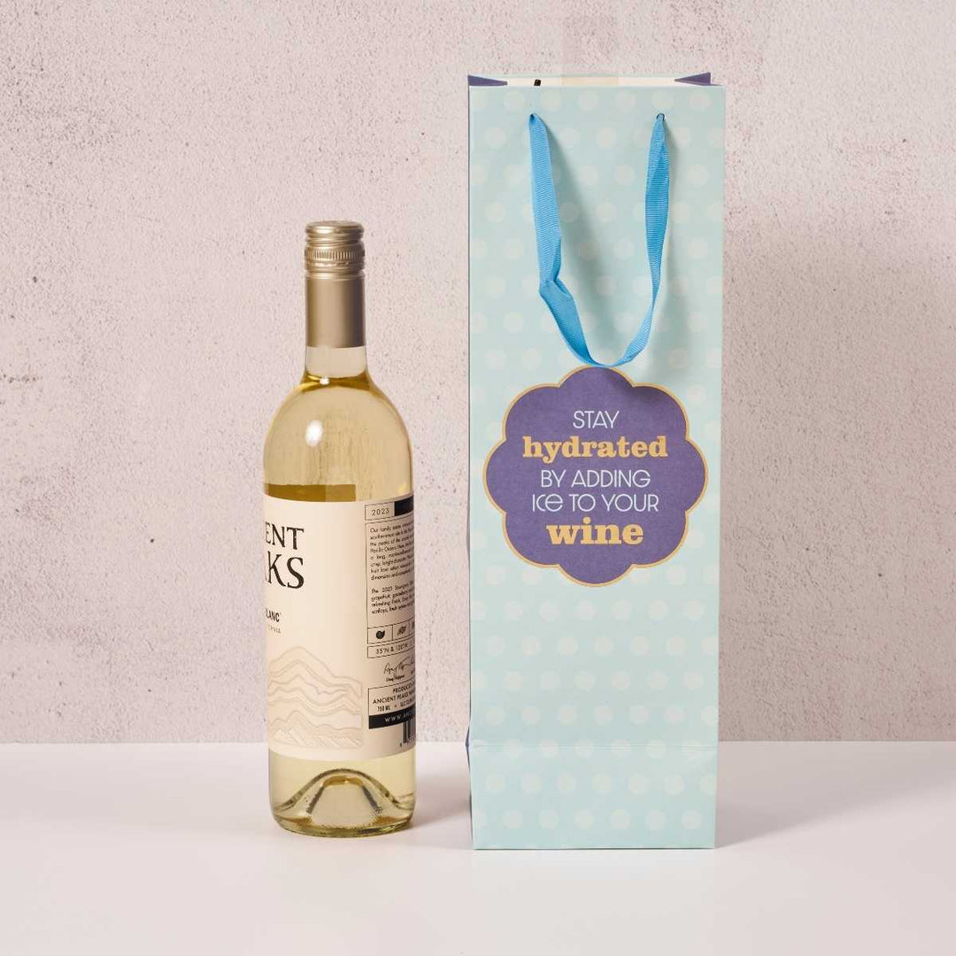 A bottle of wine stands beside the Drinks On Me Stay Hydrated Wine Bag, featuring blue polka dots, ribbon handles, and the words “Stay hydrated by adding ice to your wine”—a fun choice for hostess gifts.