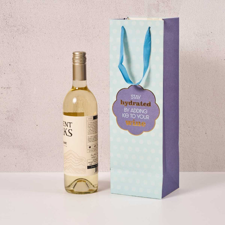 A bottle of white wine stands next to the Drinks On Me Stay Hydrated-Wine Bag, featuring blue handles and a polka dot design with “Stay hydrated by adding ice to your wine” in gold and blue—perfect for hostess gifts.