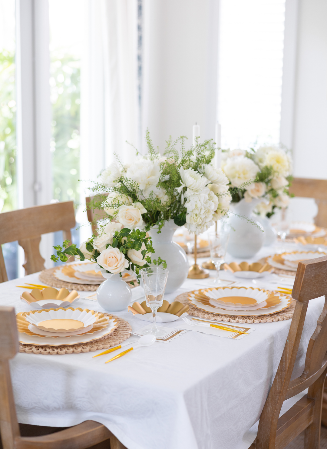 The bright room with large windows showcases the Gold & White Table Setting by Sophistiplate LLC on a table for four, featuring wooden chairs, a white tablecloth, elegant white floral centerpieces, and yellow napkins.