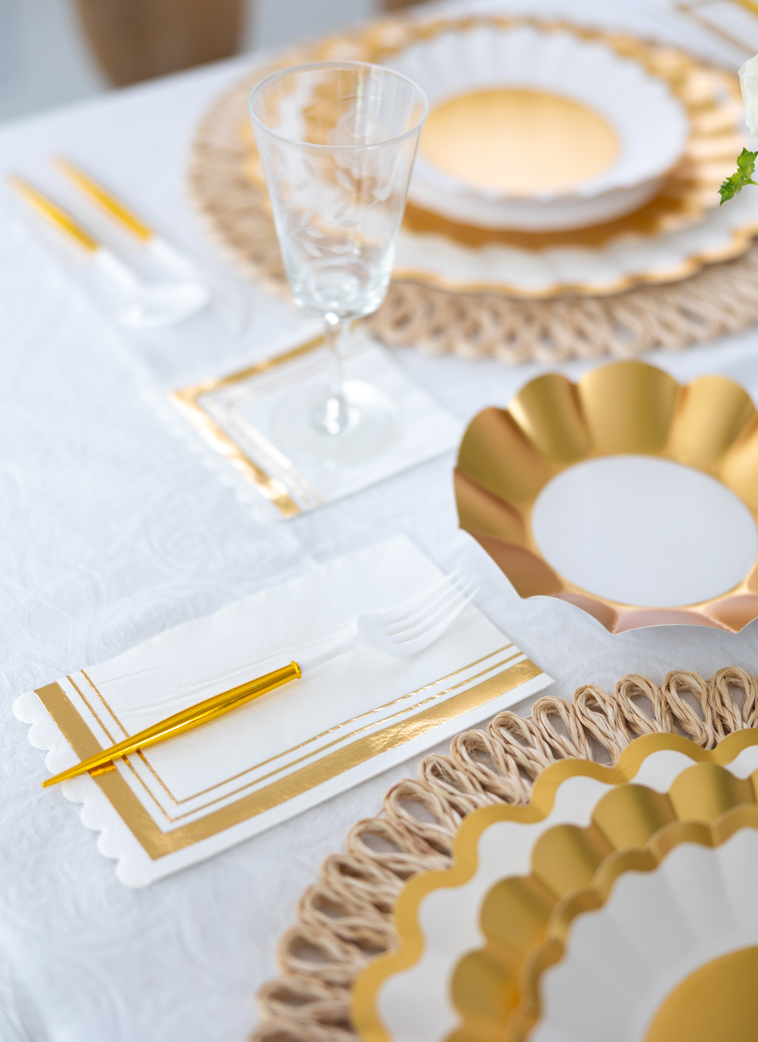 Create a stunning table with the Gold & White Table Setting by Sophistiplate LLC—featuring designer disposable plates, gold cutlery, clear wine glass, and gold-accented napkins atop a woven placemat and white tablecloth.
