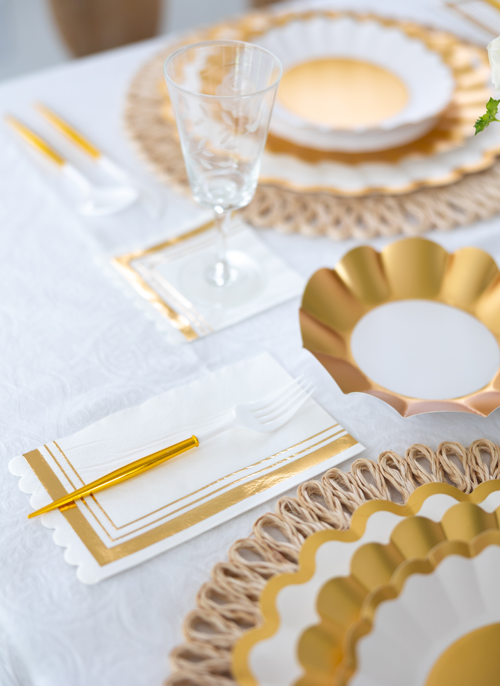 Create a stunning table with the Gold & White Table Setting by Sophistiplate LLC—featuring designer disposable plates, gold cutlery, clear wine glass, and gold-accented napkins atop a woven placemat and white tablecloth.