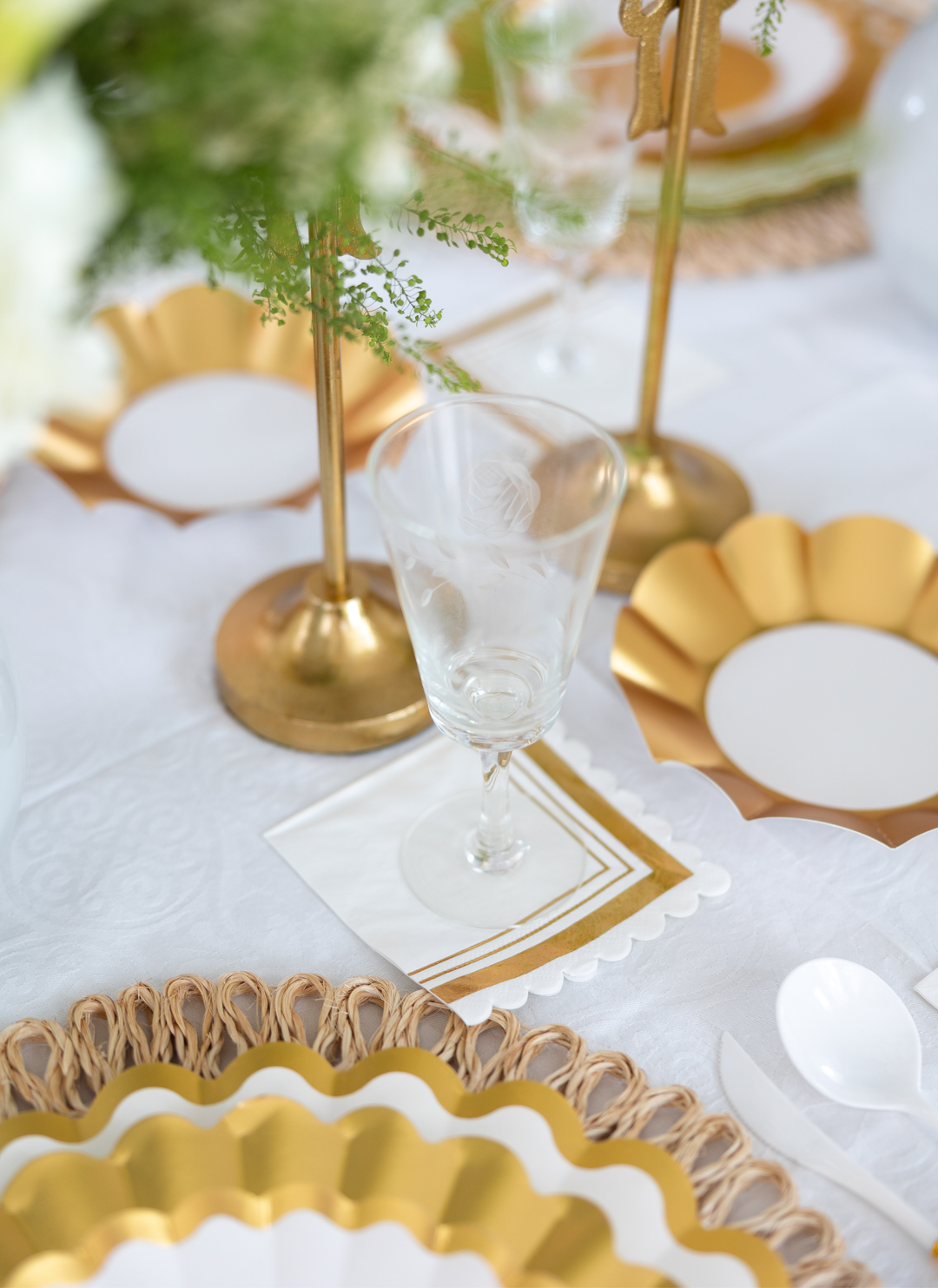 A close-up of the Gold & White Table Setting by Sophistiplate LLC showcases designer disposable plates, a clear wine glass on a gold-bordered napkin, gold candlesticks, and decorative greenery atop a white tablecloth.