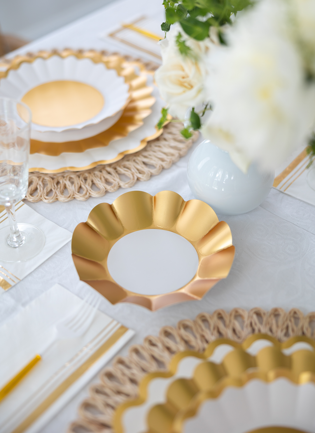 Create an elegant table with the Gold & White Table Setting by Sophistiplate LLC, featuring designer disposable plates, woven placemats, clear glassware, gold utensils, and a white vase of flowers on a white tablecloth.