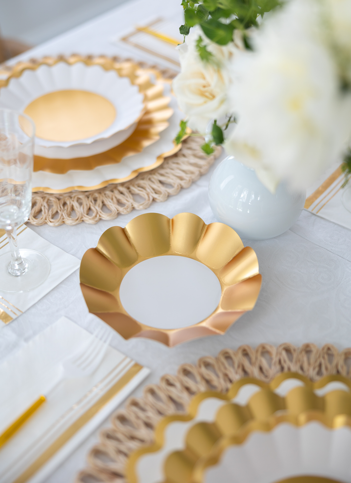 Create an elegant table with the Gold & White Table Setting by Sophistiplate LLC, featuring designer disposable plates, woven placemats, clear glassware, gold utensils, and a white vase of flowers on a white tablecloth.