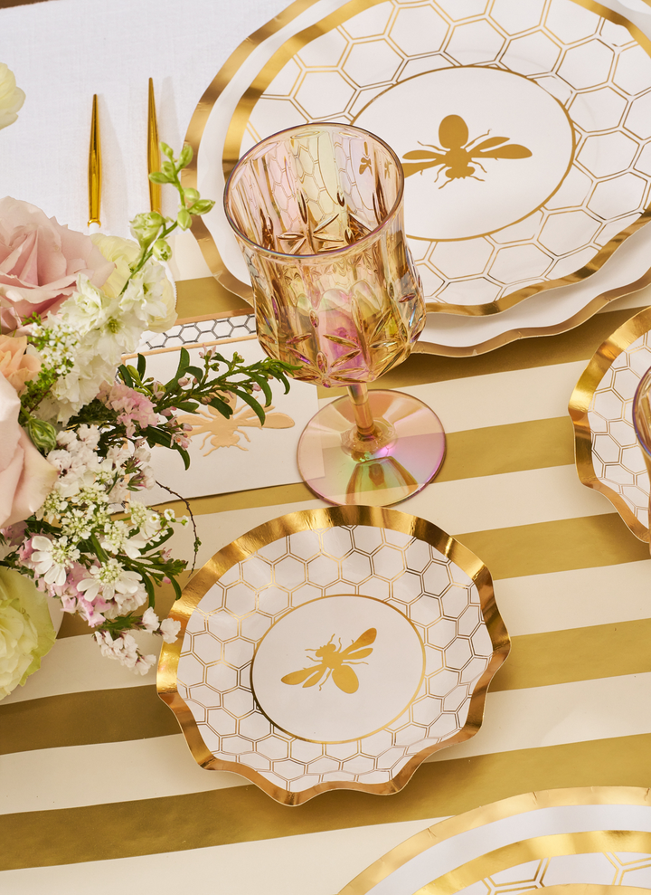 Create an elegant table setting with the Sophistiplate White Honeybee Table Setting, featuring gold-edged paper plates adorned with bee designs, a pink-tinted glass, gold utensils, and a floral arrangement atop a gold and white striped tablecloth.