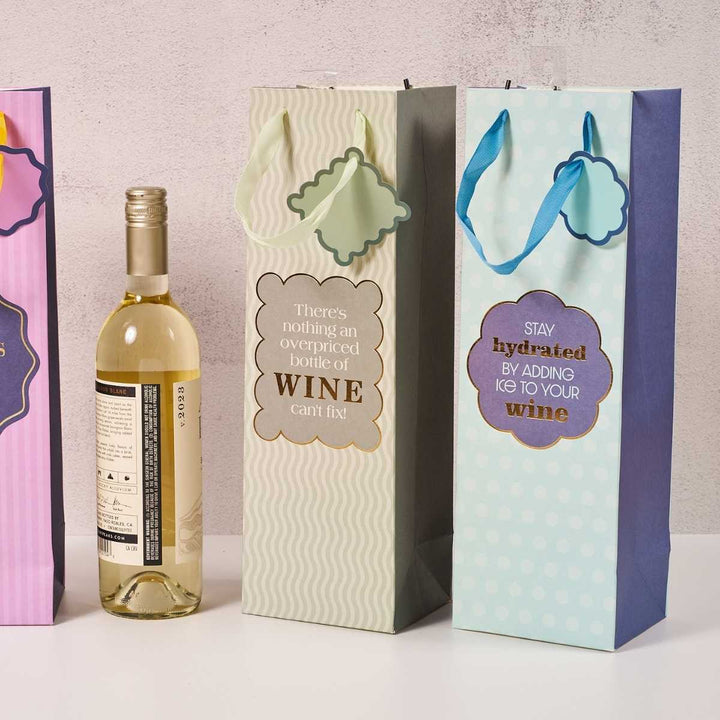 A bottle of white wine stands beside two reusable Drinks On Me "Stay Hydrated- Wine Bag" gifts—one reads, "There's nothing an overpriced bottle of wine can't fix!" and the other, "Stay hydrated by adding ice to your wine.