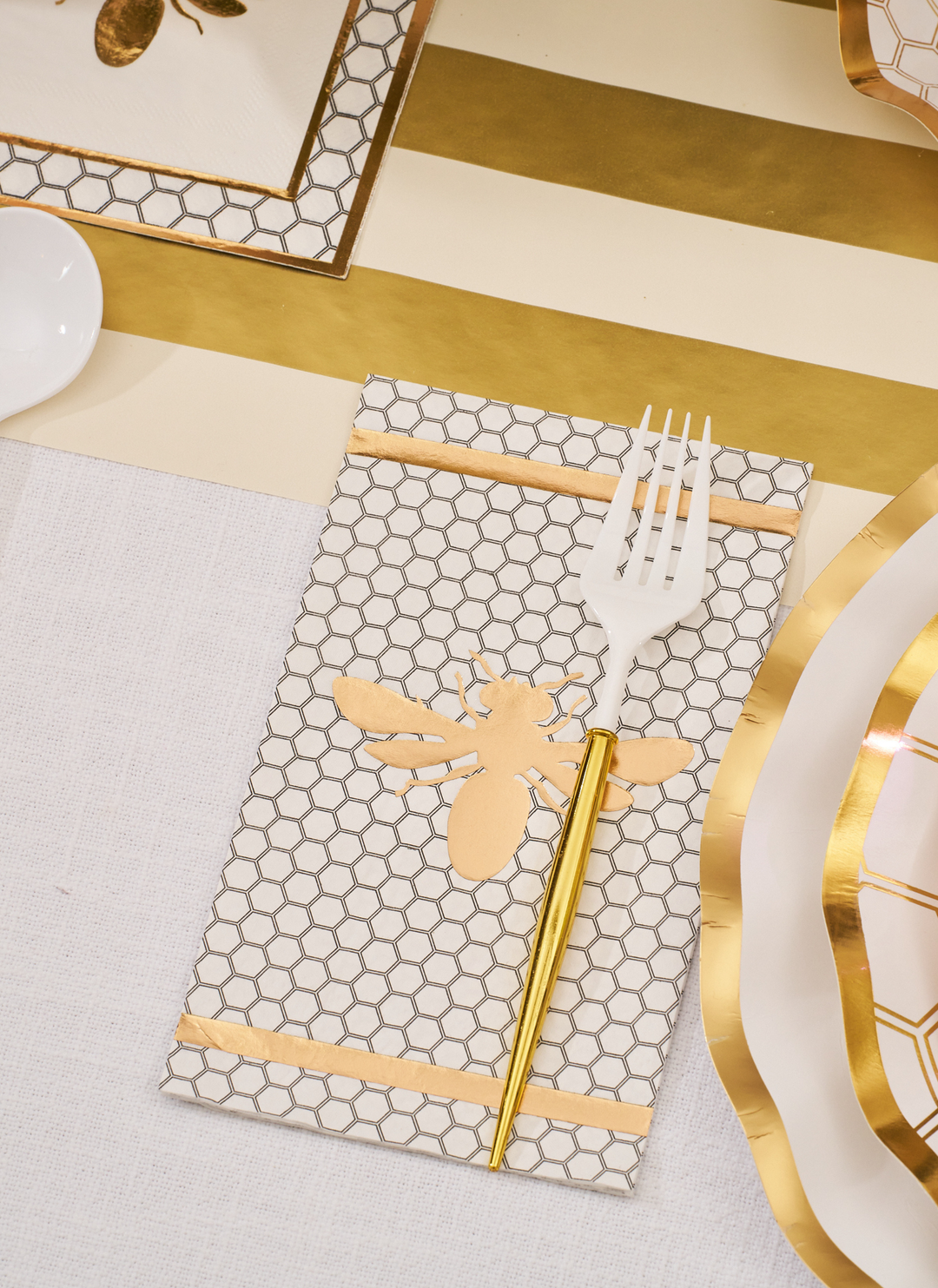 The White Honeybee Table Setting by Sophistiplate features a white fork with a gold handle set on a napkin adorned with a honeycomb pattern and gold bee design, surrounded by elegant disposable dinnerware and gold-rimmed paper plates, all arranged on a striped gold and white tablecloth.