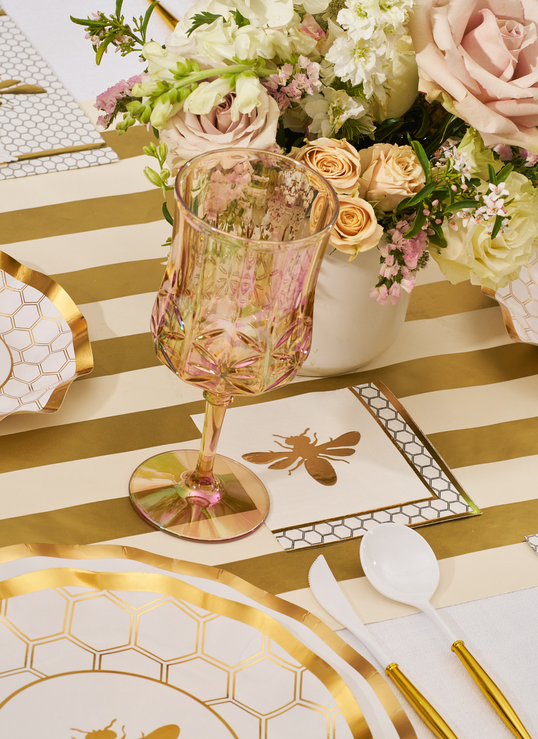 The White Honeybee Table Setting by Sophistiplate features a pink crystal goblet, gold and white striped tablecloth, elegant gold and white party plates, bee-themed napkins, and a floral centerpiece with cream and blush blooms.