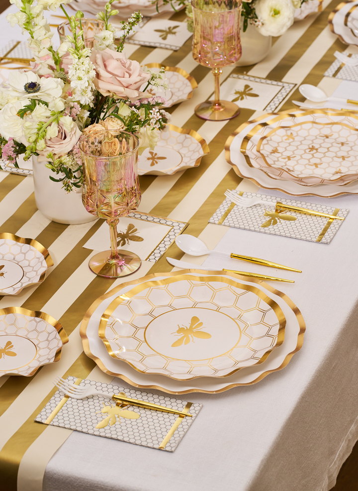 Experience the White Honeybee Table Setting by Sophistiplate: an elegant arrangement featuring gold and white party plates, gold utensils, pink glasses, and floral centerpieces atop a white tablecloth with gold-striped runners. These stylish paper plates from Sophistiplate blend seamlessly with the sophisticated décor, providing effortless cleanup without sacrificing style.