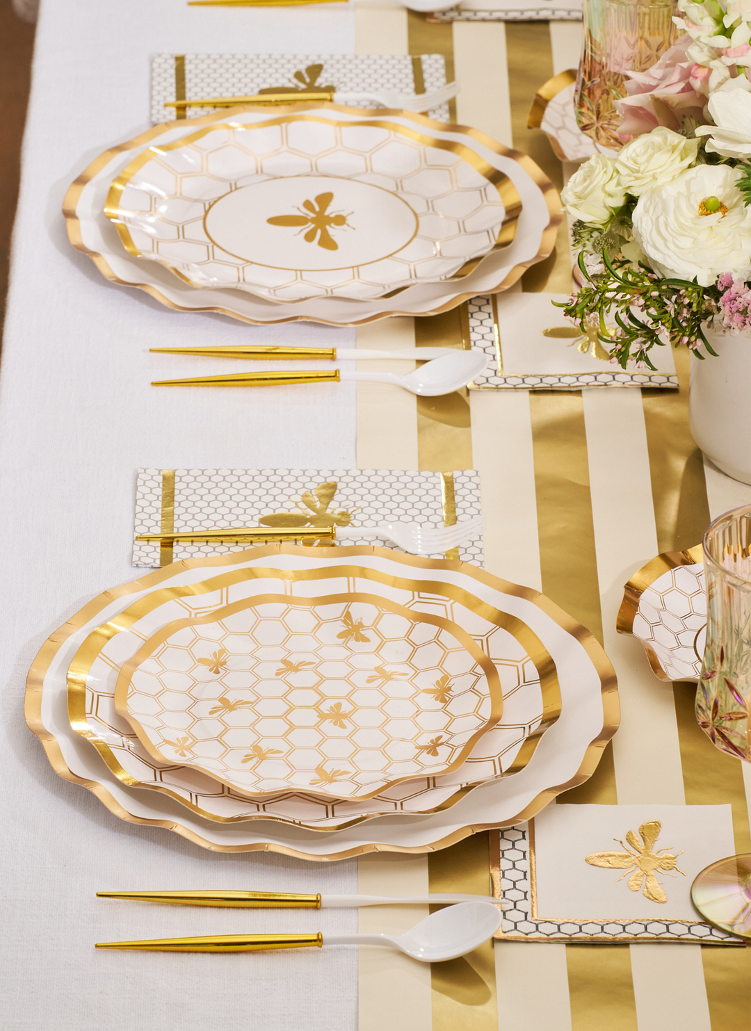 Create an elegant table setting with the Sophistiplate White Honeybee Table Setting, featuring gold and white party plates adorned with honeycomb and bee designs. Complete the look with gold flatware, a striped table runner, and a floral centerpiece of white and pale pink flowers.