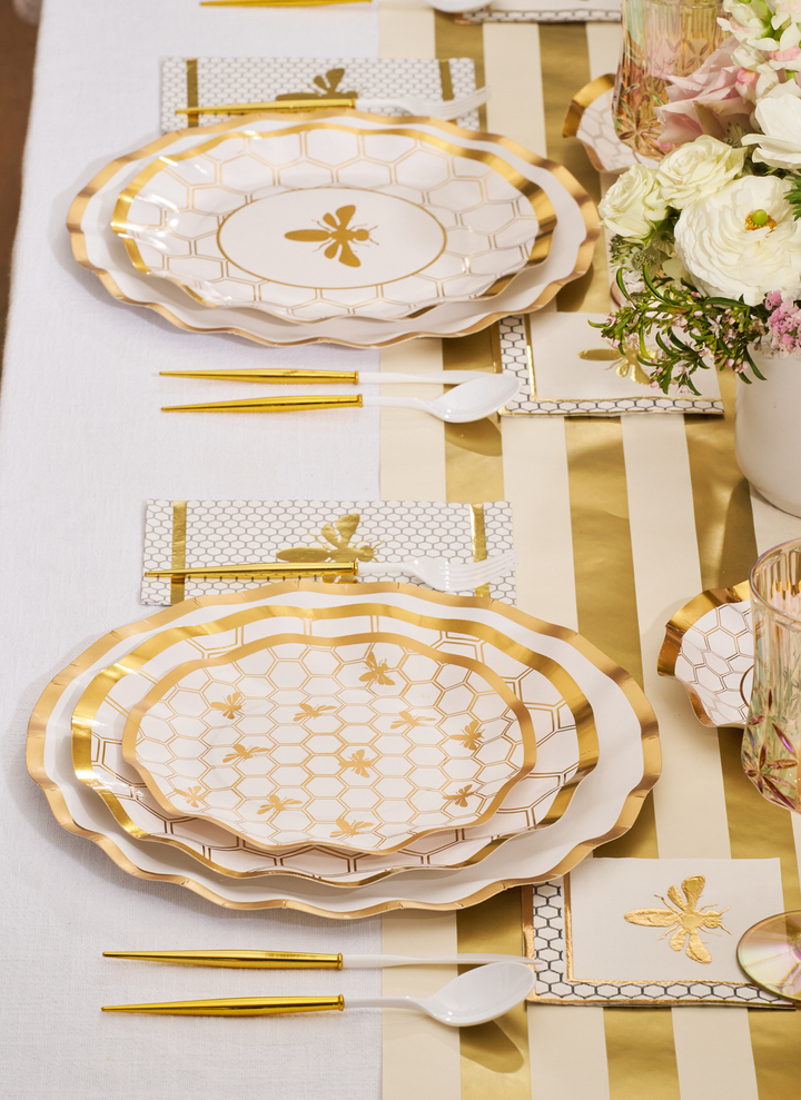 Create an elegant table setting with the Sophistiplate White Honeybee Table Setting, featuring gold and white party plates adorned with honeycomb and bee designs. Complete the look with gold flatware, a striped table runner, and a floral centerpiece of white and pale pink flowers.