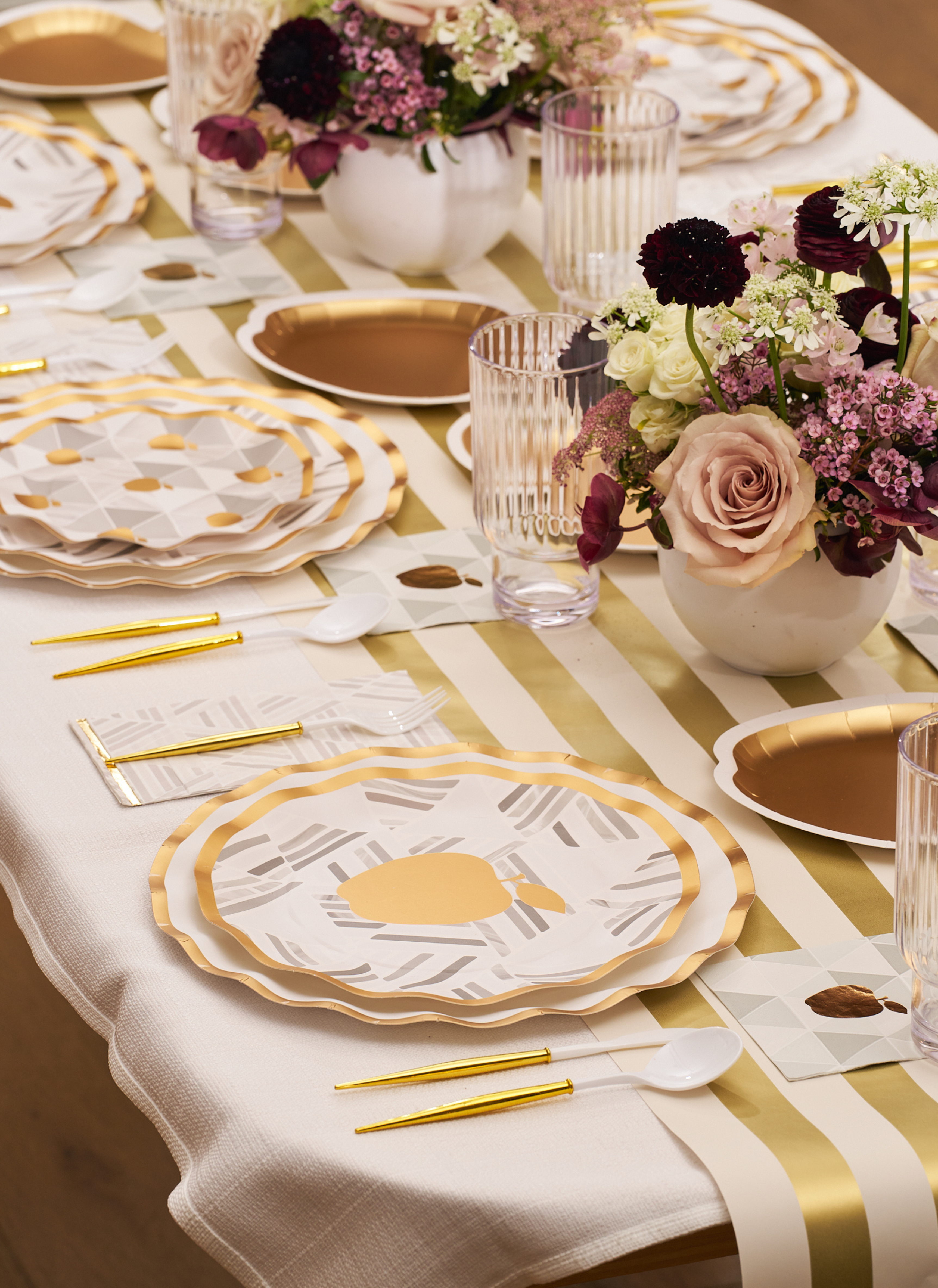 A sophisticated and inviting table is set with Sophistiplate’s Golden Apple Table Setting: gold-rimmed plates, gold cutlery, a striped runner, clear grooved glasses, and floral arrangements featuring pink and purple blooms—perfect for Rosh Hashanah or any modern gathering.