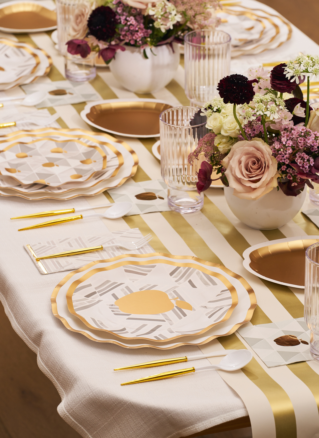 A sophisticated and inviting table is set with Sophistiplate’s Golden Apple Table Setting: gold-rimmed plates, gold cutlery, a striped runner, clear grooved glasses, and floral arrangements featuring pink and purple blooms—perfect for Rosh Hashanah or any modern gathering.