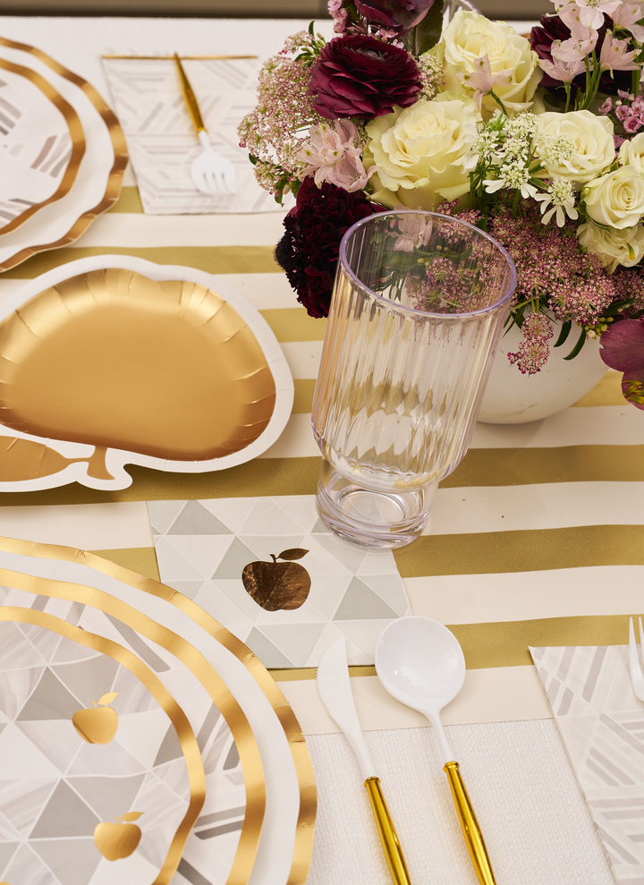 The Sophistiplate Golden Apple Table Setting includes elegant gold apple-shaped disposable plates, stylish striped placemats, a clear glass, and plastic cutlery with chic gold handles. Complemented by cheerful gold apple napkins and a beautiful centerpiece of white and purple flowers, this festive arrangement is the perfect way to celebrate Rosh Hashanah in style.