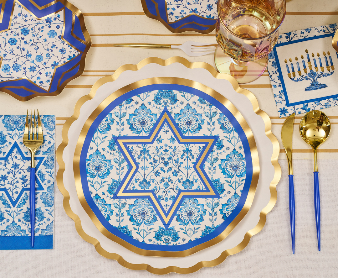 A festive tablescape is beautifully arranged with Sophistiplate’s Hanukkah Toile Table Setting, featuring a decorative plate adorned with a blue and gold Star of David pattern. The elegant scene is complemented by a gold fork, spoon, and knife, a blue floral napkin, a menorah-themed napkin, and a festive drink. These stylish disposable plates from Sophistiplate blend elegance and convenience for your Hanukkah celebration.