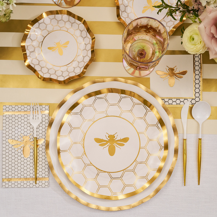 A table setting features Sophistiplate’s Honeybee White Wavy Paper Dinner Plates (8pk), gold and white plates with bee motifs, gold-rimmed glassware, coordinating utensils and napkins, plus a sparkling gold bee centerpiece on a striped tablecloth.