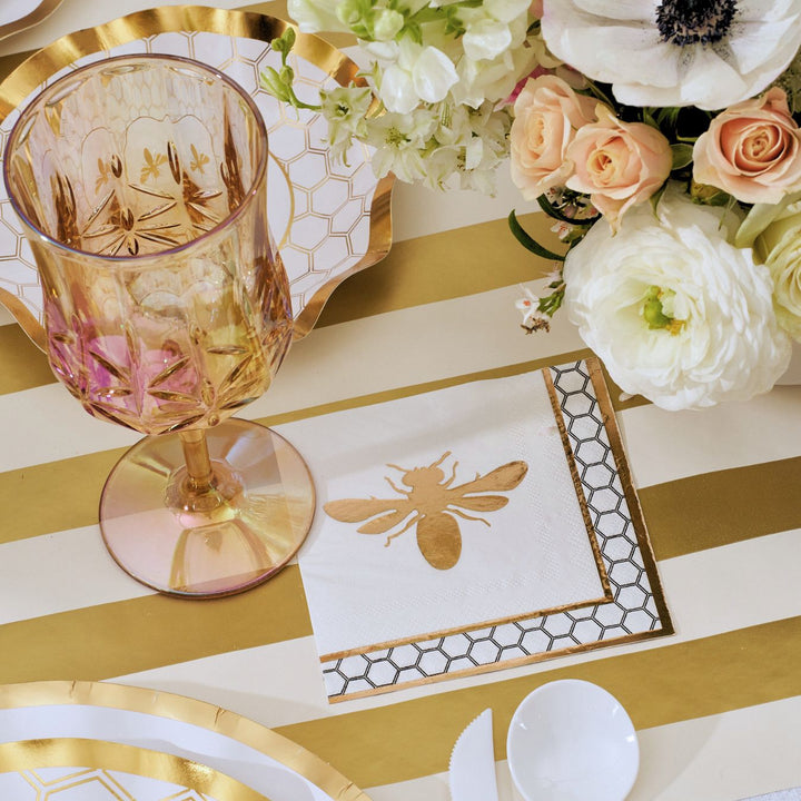 The White Honeybee Table Setting by Sophistiplate features a gold and white striped tablecloth, pink-tinted glassware, a floral centerpiece, honeycomb-patterned plates, and napkins embellished with a gold bee design. Elegant party tableware—including white plastic cutlery and gold-rimmed paper plates—adds a refined yet convenient finishing touch to the setting.