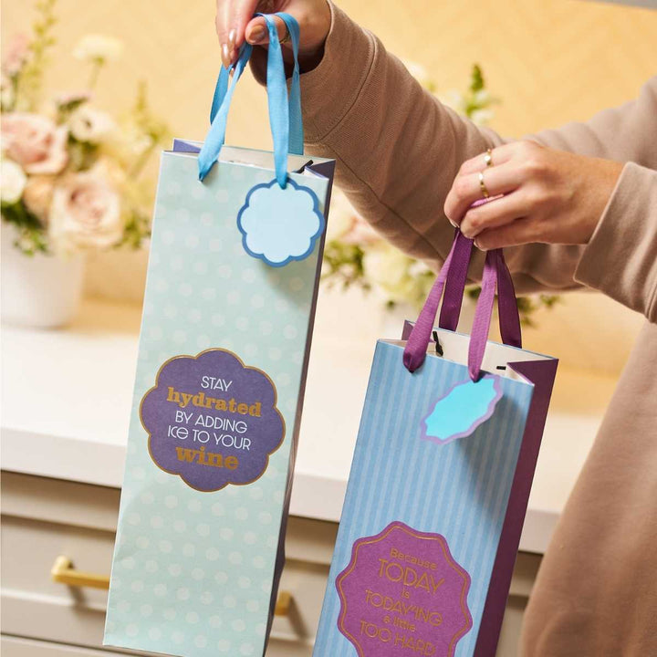 Two hands hold Drinks On Me Stay Hydrated wine bags, each with ribbons and tags—great for hostess gifts. The blue reusable bag says, "Stay hydrated by adding ice to your wine," while the purple one reads, "Because today is definitely too hard.