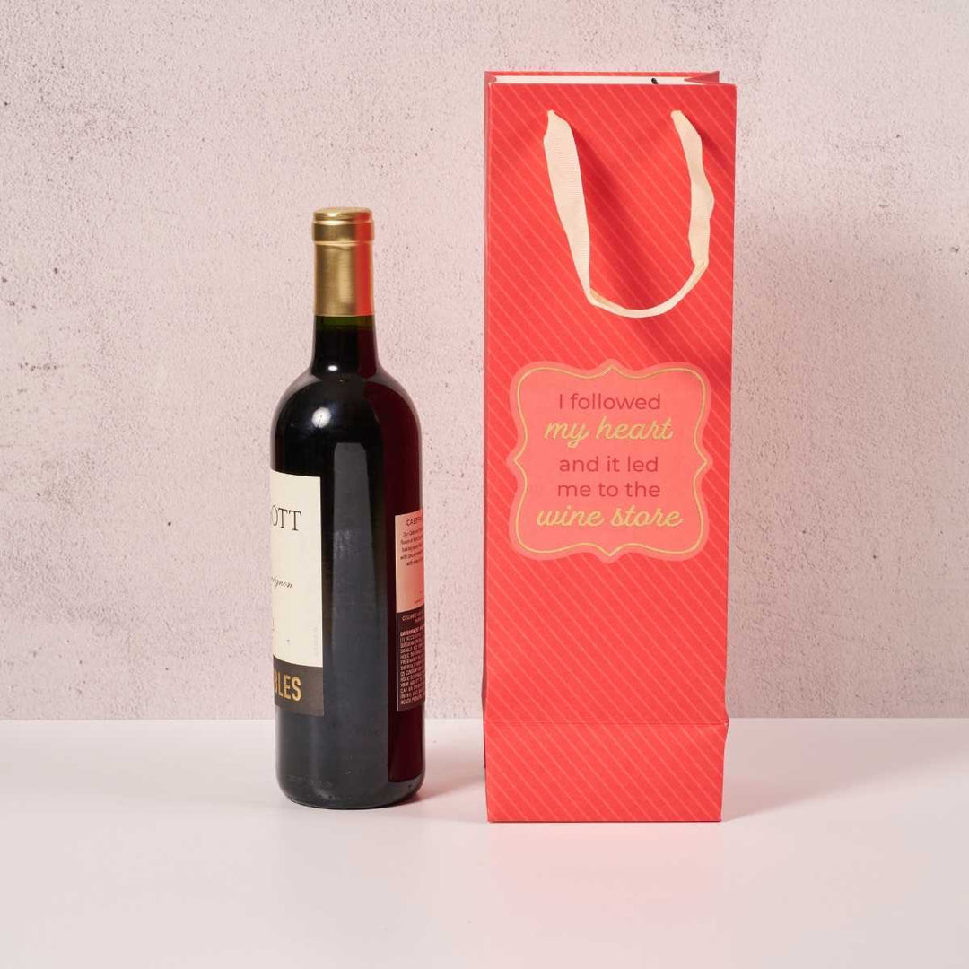A bottle of red wine is beside the Drinks On Me Wine Store- Wine Bag, featuring white handles and the phrase "I followed my heart and it led me to the wine store.