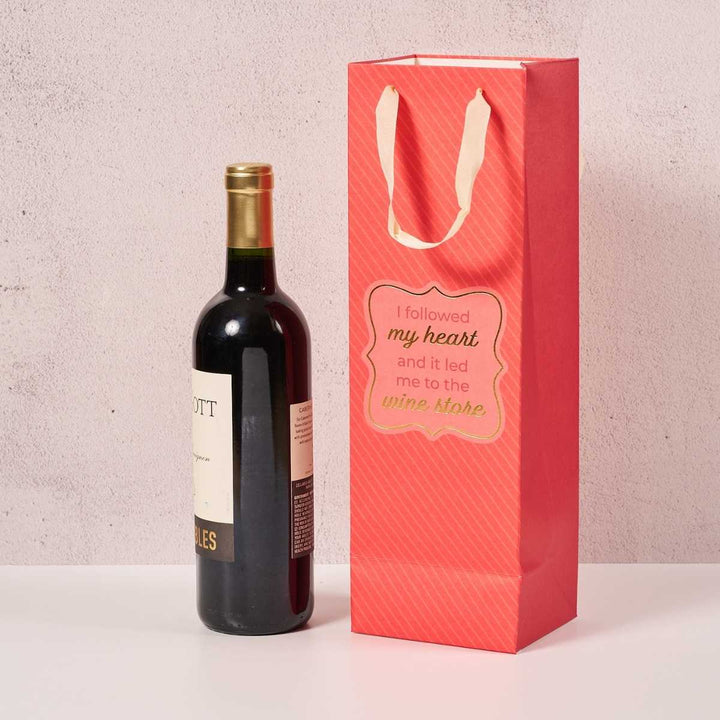 A bottle of red wine stands next to the Drinks On Me Wine Store- Wine Bag, a pink bag with cream handles and a label that reads, "I followed my heart and it led me to the wine store.