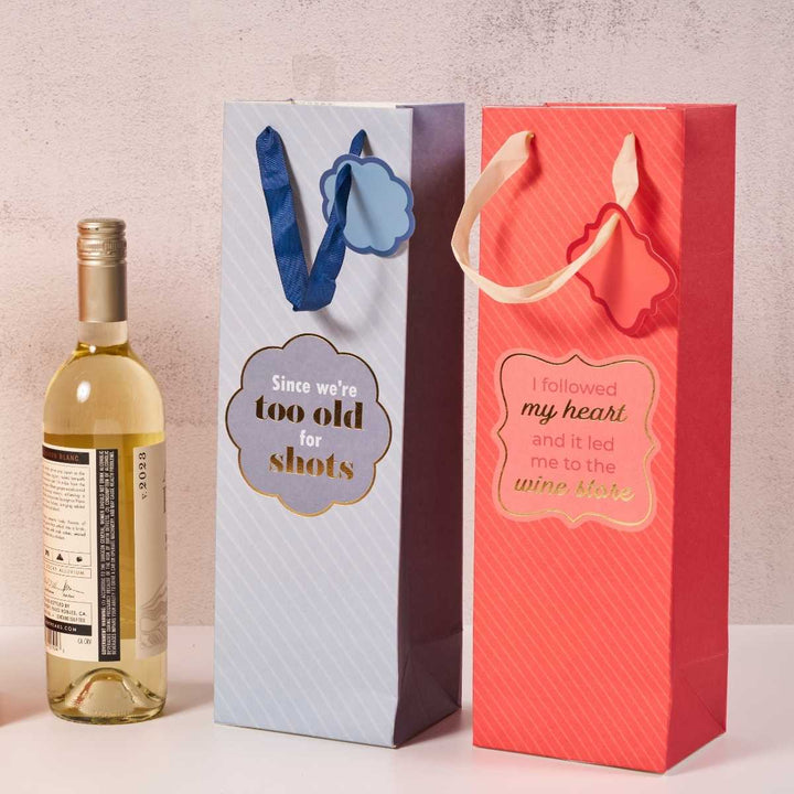Two funny reusable wine bags from Drinks On Me: the blue “Since we're too old for shots” and the red Wine Store- Wine Bag “I followed my heart and it led me to the wine store,” plus a bottle of white wine.