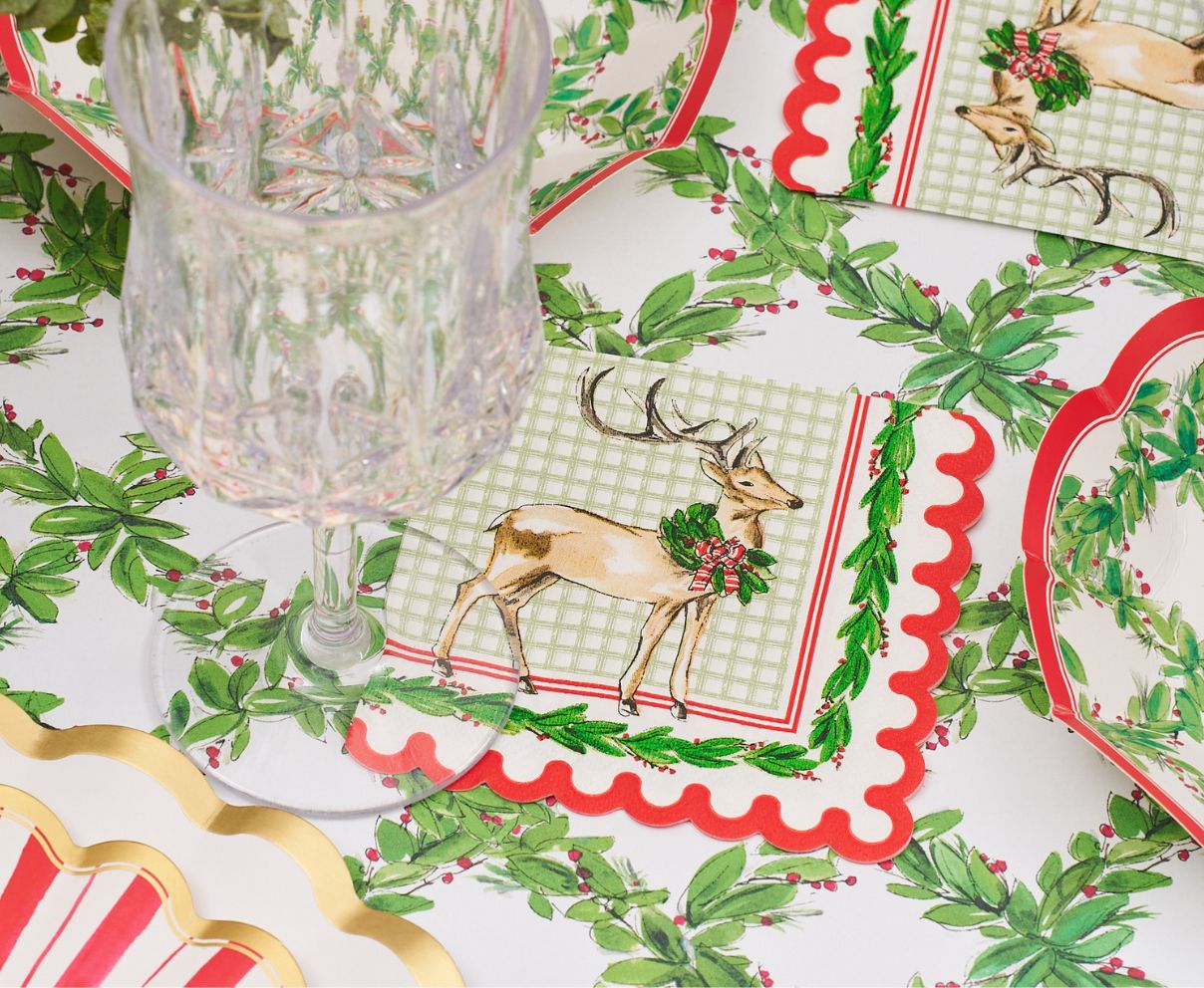 Create a charming Christmas table with a clear glass, plates, and napkins adorned with a reindeer and festive foliage, all set on the Rosanne Beck Holly Berry Trellis Table Runner (20x12