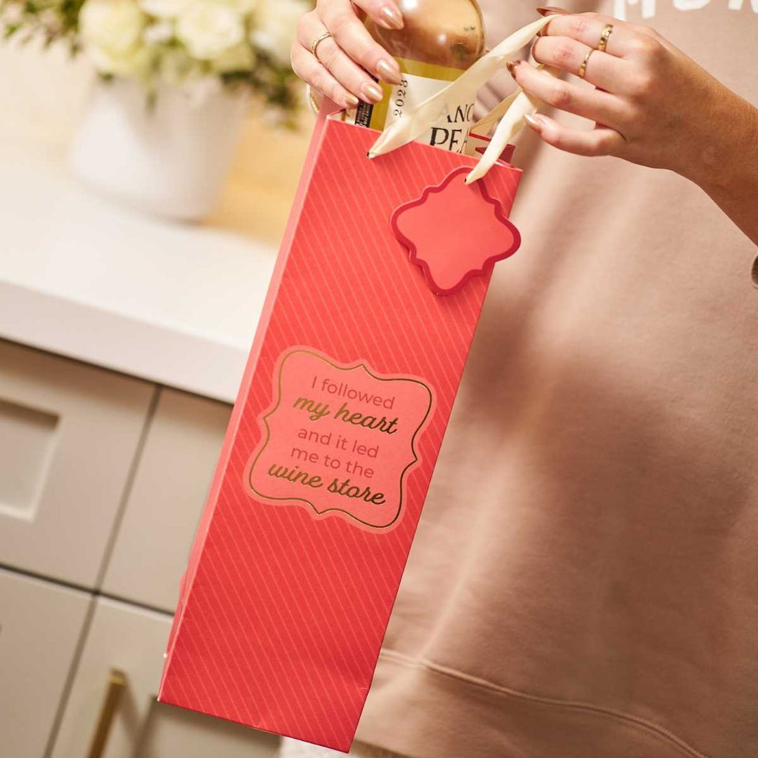 A person in a kitchen holds the "Wine Store- Wine Bag" by Drinks On Me, which says, "I followed my heart and it led me to the wine store," with a wine bottle inside and flowers and white cabinets in the background.