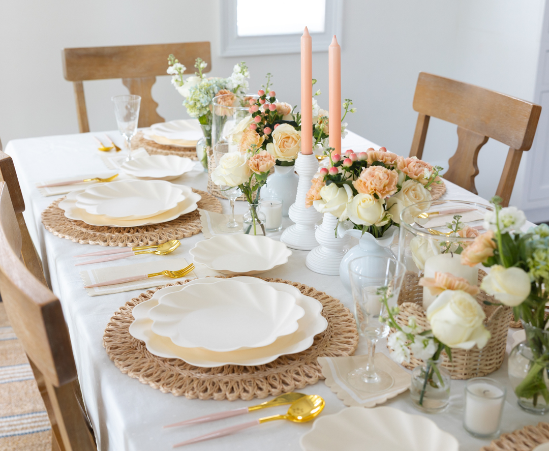 A dining table set with Sophistiplate's Simply Eco Dinner Plate-Cream (8/pkg), on woven placemats, gold utensils, pastel floral centerpieces, peach candles, and clear glasses. Wooden chairs complete the elegant, inviting setting.