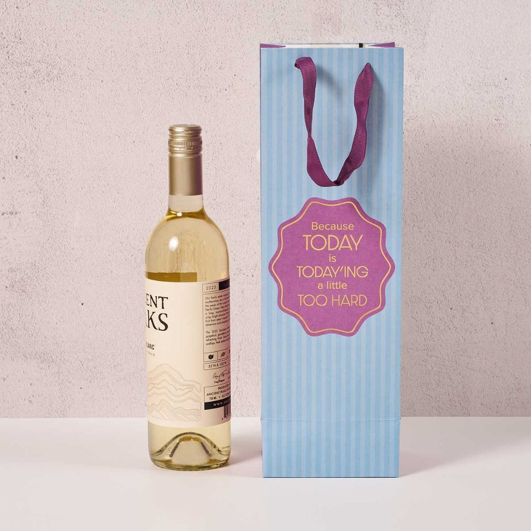 A bottle of white wine stands beside the Drinks On Me Today'ing-Wine Bag, a light blue striped reusable bag with purple handles and a label that says, "Because today is today-ing a little too hard.
