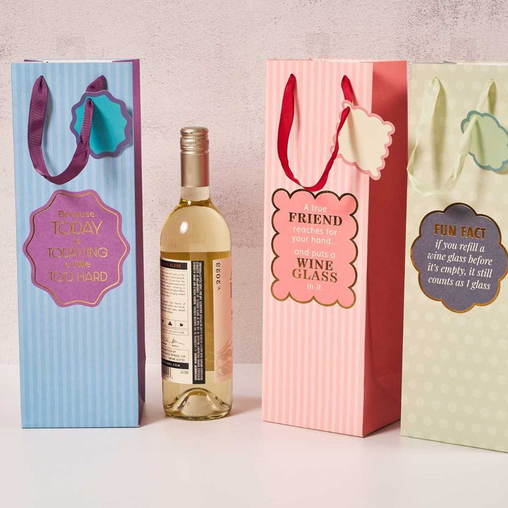 Three Today'ing-Wine Bags from Drinks On Me, featuring colorful patterns and witty wine quotes, are displayed beside a white wine bottle. With attached tags, these reusable bags make the perfect hostess gift.